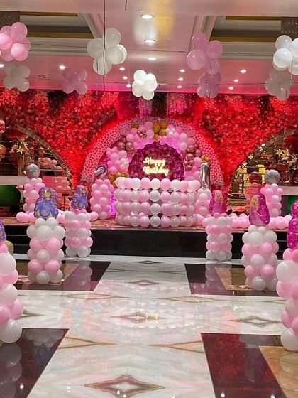 A grand princess-themed hall decoration. The setup includes a stage with a red floral backdrop, pink and white balloon columns topped with character foils, and a balloon-covered cake table.