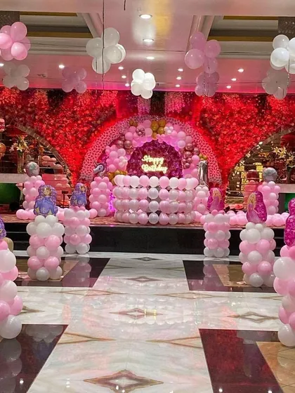 A grand princess-themed hall decoration. The setup includes a stage with a red floral backdrop, pink and white balloon columns topped with character foils, and a balloon-covered cake table.