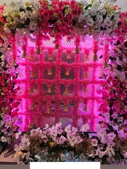 This decor combines a lattice of pink flowers with golden bells, lit up with pink lighting for a vibrant and celebratory feel.