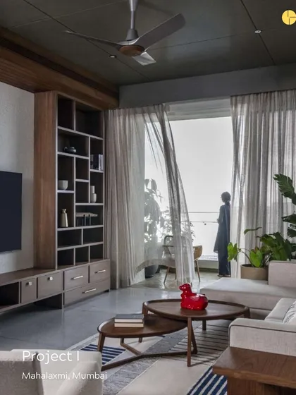 A glimpse into 'Project I' in Mahalakshmi, Mumbai. The living room is designed to feel grounded and authentic, with a custom wooden bookshelf, comfortable seating, and sheer curtains that filter the city light, creating a soft, pleasant atmosphere.
