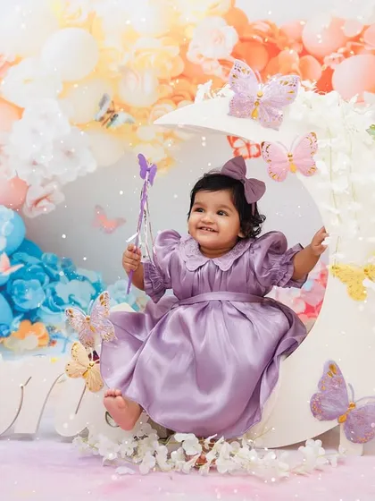 A little princess sits on a moon prop, surrounded by pastel balloons and butterflies for her first birthday.