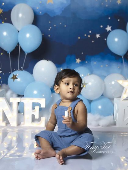 A classic first birthday portrait before the cake smash begins. The baby is dressed in a cute blue outfit, ready for his big celebration against the starry sky backdrop.