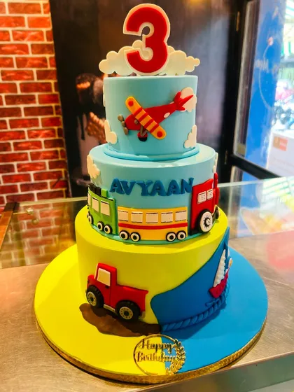 A three-tier transportation-themed cake for a 3rd birthday, featuring a tractor, train, bus, and airplane.