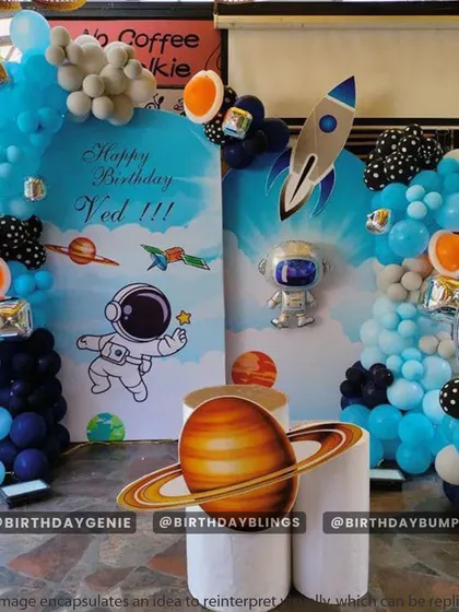 This outdoor space party features large planet cutouts, including Saturn on a plinth, and a fun astronaut character. The blue and black balloons with pops of orange create a striking look.