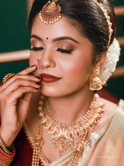 A classic beauty shot focusing on the flawless makeup and traditional gold jewelry. The bride's pose, gently touching her nose ring, is a timeless gesture of adornment.