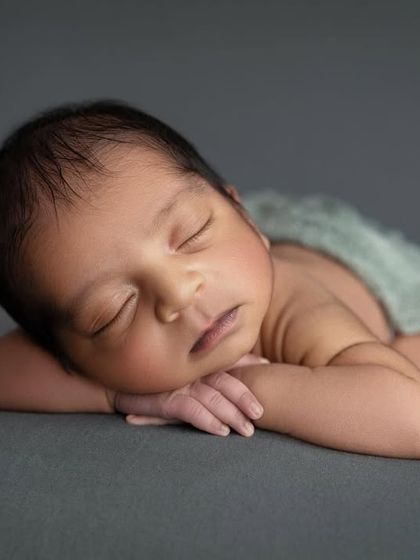A beautiful close up of a newborn in the 'chin on hands' pose against a solid grey backdrop. The lighting perfectly highlights the contours of the baby's face.