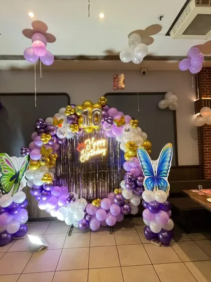 A butterfly-themed ring decoration inside a restaurant, using purple, white, and gold balloons, with large butterfly cutouts and a 'Happy Birthday' neon sign.