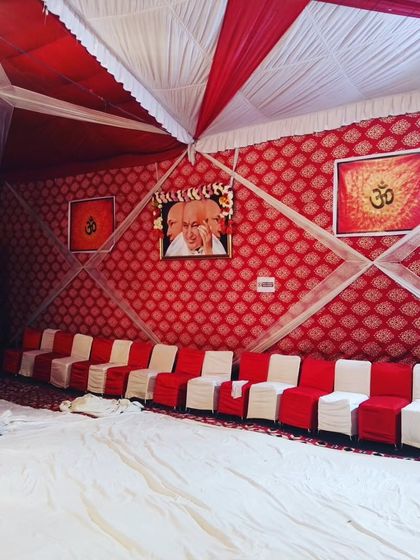 This is a simple yet respectful setup for a Guruji satsang inside a tent. The walls are covered with patterned red fabric, and a dedicated space is created with portraits and floor seating.