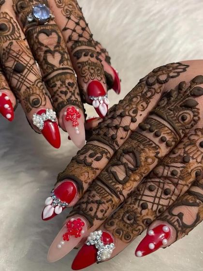 A bold and beautiful bridal nail design. This set features vibrant red nails with pearl dots, intricate 3D floral and bow embellishments, and sparkling rhinestone accents, all designed to stand out alongside the bridal mehendi.