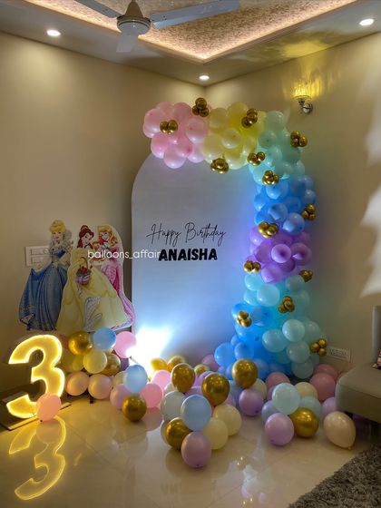 A dreamy Disney Princess theme for a 3rd birthday. This setup includes a pastel rainbow balloon arch, a large light-up number '3', and cutouts of Cinderella, Belle, and Aurora.