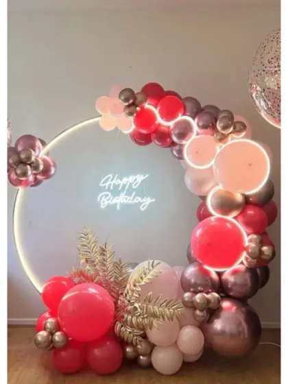 A modern and chic birthday backdrop perfect for photos. This setup includes a lit-up circular frame, a custom neon "Happy Birthday" sign, and a stylish balloon garland in shades of red, pink, and rose gold.