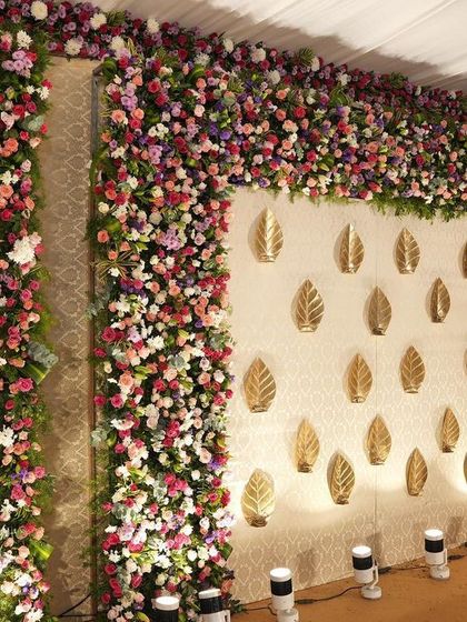 A beautiful floral wall with columns of vibrant pink, purple, and white flowers. The centerpiece is a cream panel adorned with illuminated golden leaf motifs.