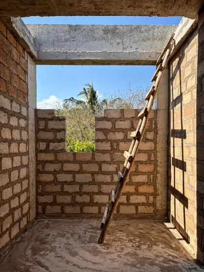 A simple wooden ladder leans against a wall in a room under construction at Kizikula. The image captures the raw, handmade quality of the building process, with sunlight streaming through the open roof.