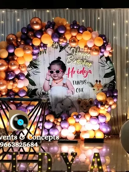 A stylish first birthday decor with a personalized photo backdrop of the birthday girl. The metallic balloon arch in purple and orange, along with illuminated 'ONE' letters, creates a chic and modern look.