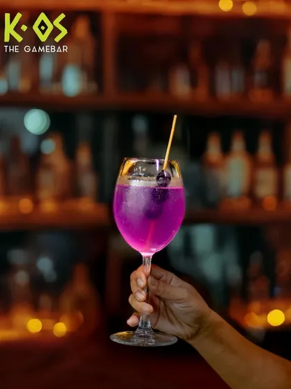 This drink just entered its main character era. A vibrant purple cocktail held up against the backdrop of our well-stocked bar.