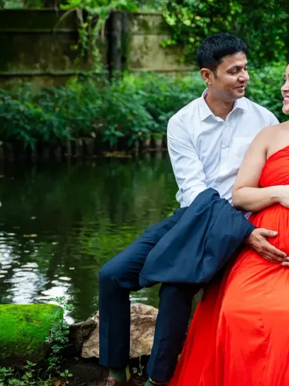 A quiet, candid moment shared between an expecting couple by the water. The vibrant red of her dress stands out beautifully against the serene, green landscape of the park.