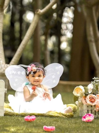 An enchanting outdoor butterfly-themed cake smash. The baby girl, dressed as a fairy with tiny wings, enjoys her first taste of cake in a beautiful garden setting, creating a magical and whimsical portrait.