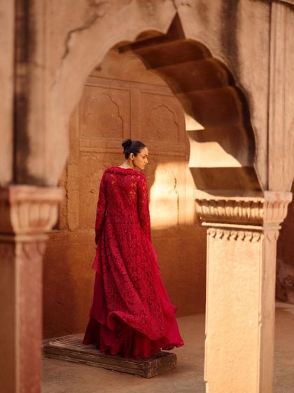 A single model styled in a full-length red lace jacket and skirt set. The location's archways and warm stone create a beautiful frame, highlighting the rich color and texture of the outfit.