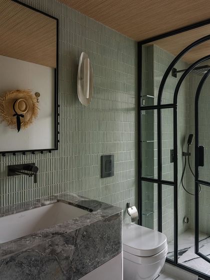 The master bathroom is a calm sanctuary, featuring sage green tiles, a marble vanity, and black metal accents. The arched shower door adds a touch of softness to the modern design.