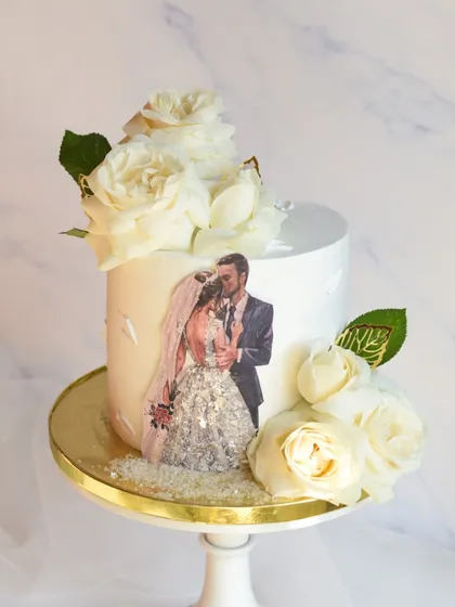 An anniversary cake that tells a personal story, featuring an edible print of the couple's wedding photo. The cake is elegantly decorated with fresh white roses and touches of edible glitter on the bride's dress.