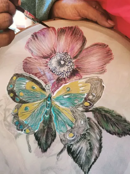 A student carefully adds detail to a butterfly and flower design during a fabric painting class. We use embroidery hoops to hold the fabric taut, allowing for precise and detailed work.