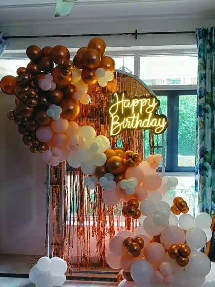 A birthday decoration with a rose gold fringe backdrop. The setup features a balloon arch in bronze, peach, and white, and a "Happy Birthday" neon sign.