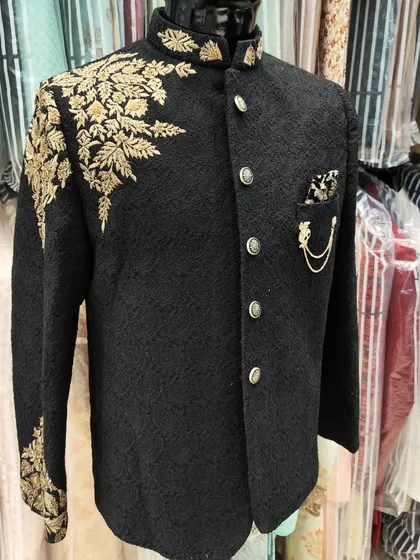 A showcase of regal craftsmanship. This black Jodhpuri suit features intricate gold hand embroidery on the shoulder and sleeve, creating a look of timeless elegance perfect for a groom or a grand event.