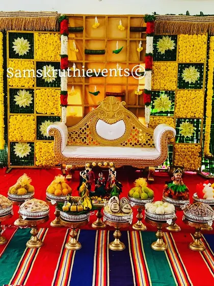 A classic baby shower setup with a checkerboard backdrop of yellow marigolds and woven green leaves. The traditional sofa and a full display of offerings on a striped mat complete this authentic South Indian look.