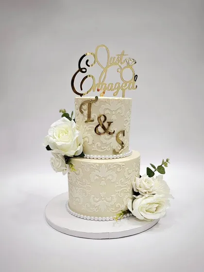 A two-tier engagement cake with intricate white-on-white stenciling, white roses, and a custom gold topper.