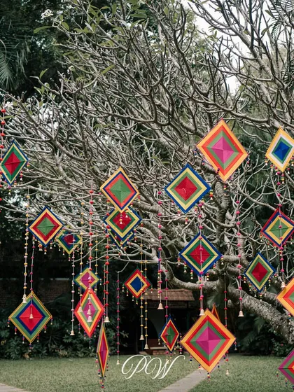 Colorful kite-like hangings and bells adorn a tree for a Mehendi ceremony, adding a playful and festive touch to the outdoor space.