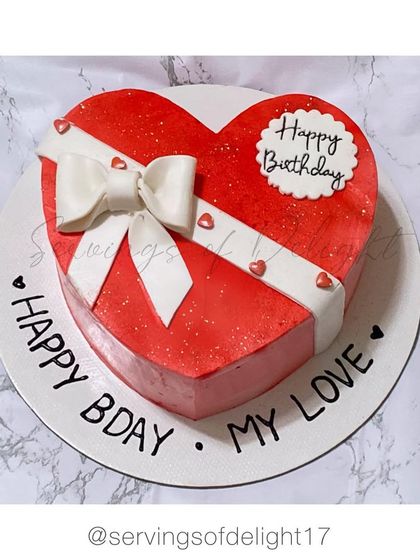 A classic red velvet heart-shaped cake designed to look like a gift box. The fondant bow and "Happy Bday My Love" message make it a truly romantic gesture.