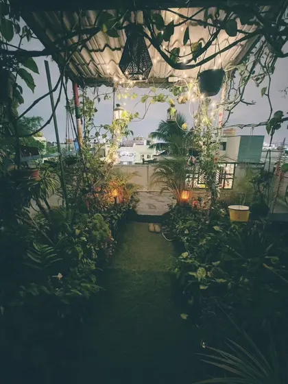 A view looking out from the garden path at night. The archway of vines and lights frames the city skyline, blending the urban environment with this natural oasis.