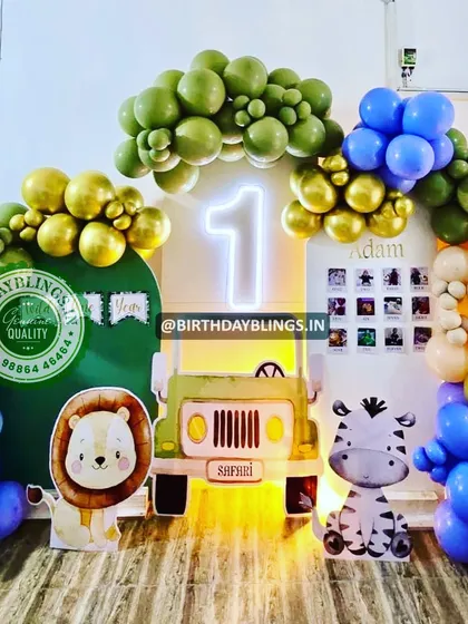 A safari jeep cutout is the star of this jungle-themed first birthday. The setup includes cute lion and zebra characters and a balloon arch in green, blue, and gold, with a neon '1' sign.