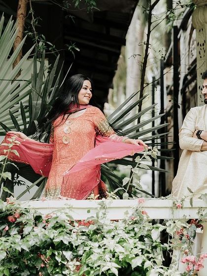 A couple in traditional attire shares a look on a balcony surrounded by lush greenery, creating a romantic and peaceful scene.