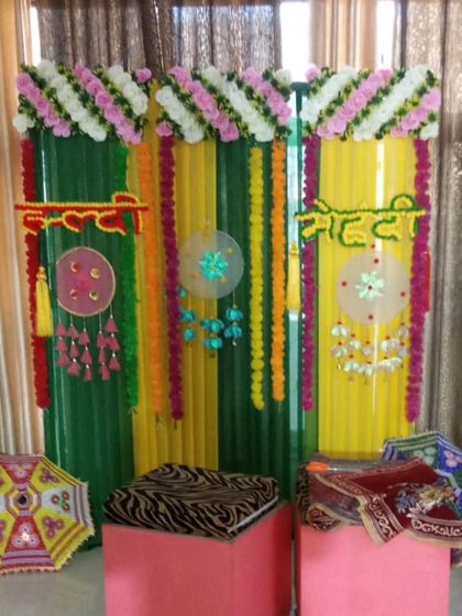 A simple and colorful backdrop for a home Haldi or Mehandi. The setup uses green and yellow drapes, marigold strings, and traditional hangings, with small stools and umbrellas as props.