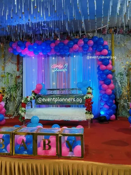 A large-scale baby shower or Dohale Jevan setup in a hall. The stage includes a decorated sofa, pink and blue balloon arch, and "BABY" boxes filled with balloons.