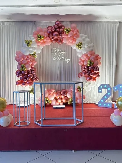 A clean and elegant 21st birthday stage setup. It features a circular balloon arch in pink, white, and gold, with a 'Happy Birthday' sign and marquee numbers.