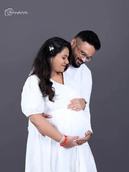 A simple and sweet couple's portrait in matching white outfits against a solid grey backdrop. The focus is purely on their connection and anticipation.
