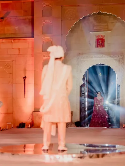 The groom waits on a mirrored platform as his bride appears in a doorway, framed by dramatic lighting. This 'first look' moment is filled with anticipation and cinematic beauty.