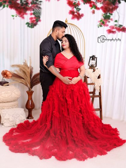 A romantic studio portrait with a bohemian theme. The couple is surrounded by pampas grass and a golden birdcage prop, with the mother-to-be in a stunning red gown.