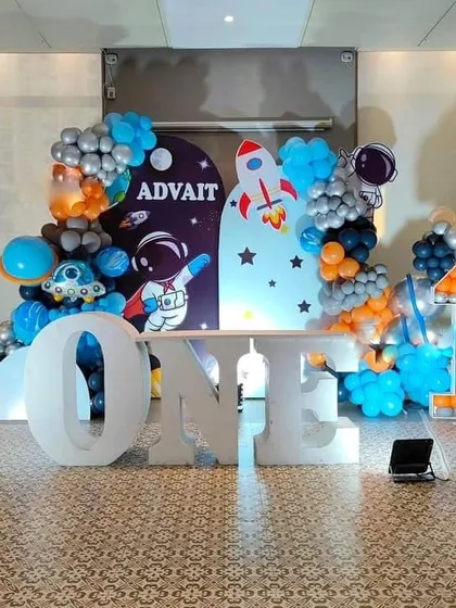 An out-of-this-world space theme for a first birthday, with large "ONE" marquee letters in the foreground. The backdrop features astronauts and rockets, framed by a blue, orange, and silver balloon arrangement.