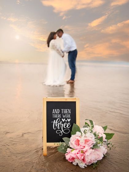A beautiful beach scene with a sign that says "And then there were three". The focus on the announcement details with the couple blurred in the background creates an artistic and emotional image.
