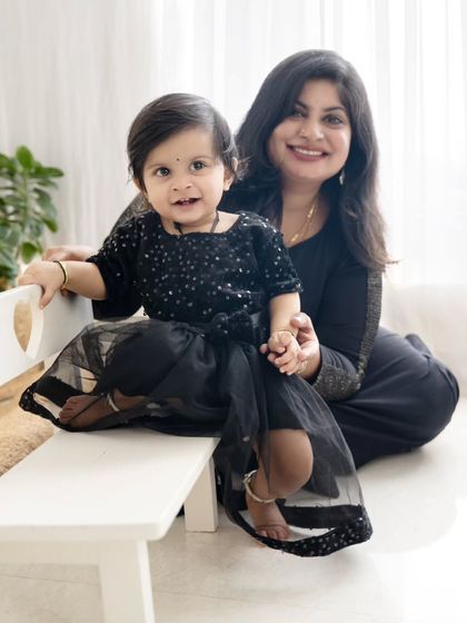 A mother sits with her daughter on a small white bench, both in matching black dresses.