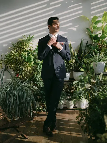 A groom adjusts his tie, surrounded by a wall of lush green plants. The natural setting and the warm sunlight create a fresh and modern groom portrait.