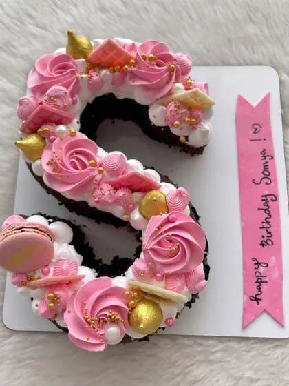 A letter "S" cake in pretty shades of pink and white, decorated with macarons, meringue kisses, and gold sprinkles.