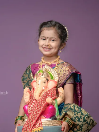 A close-up shot of the young girl in her traditional attire, holding the Ganpati idol. Her bright smile and the vibrant colors of her saree make this a joyful and captivating portrait.