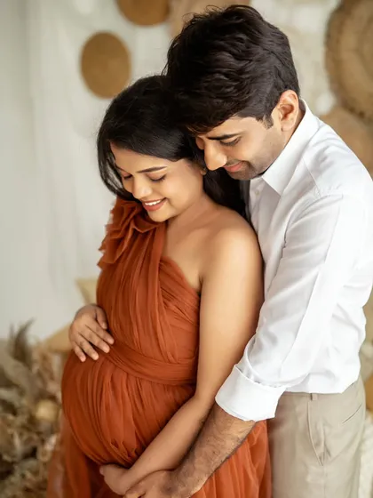 An intimate embrace between the expecting couple. The warm, earthy tones of her gown and the rustic studio set create a cozy and romantic feeling.