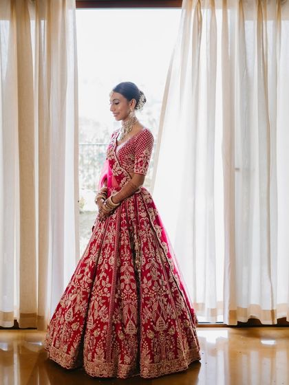 A full-length portrait of the bride by the window. The natural light beautifully highlights the radiant finish of her makeup and the intricate details of her lehenga.