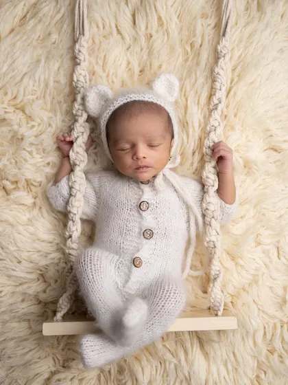 Sometimes, simple and sweet is the most beautiful. This little one is dressed in a cozy, hand-knitted teddy bear outfit, sleeping soundly on a miniature swing against a soft, textured backdrop.