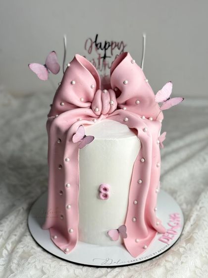 A tall white cake with a dramatic, draping pink fondant bow. The addition of small butterflies gives this birthday cake a playful and pretty finish.